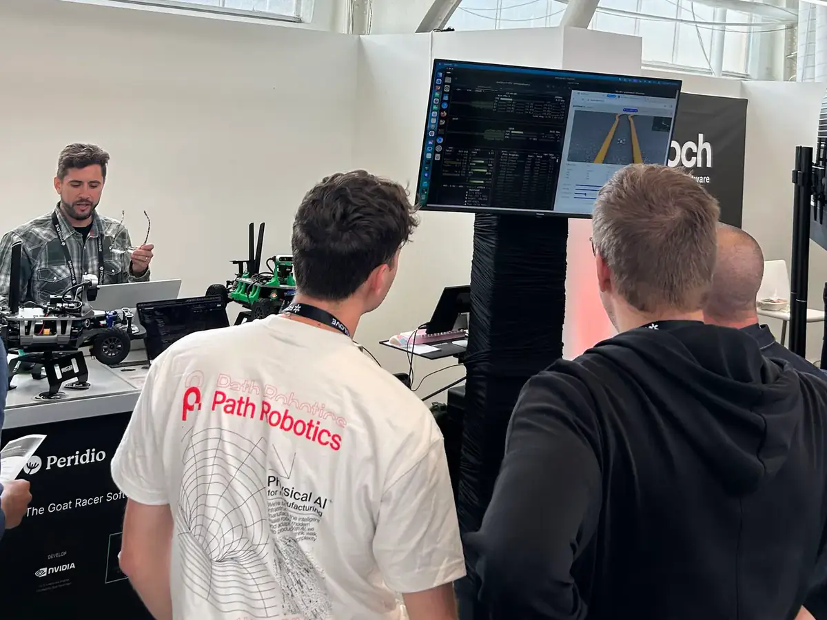 Engineers at the Peridio booth watch a large screen showing system stats beside a simulated track, with two GOAT Racer 3 cars on the counter