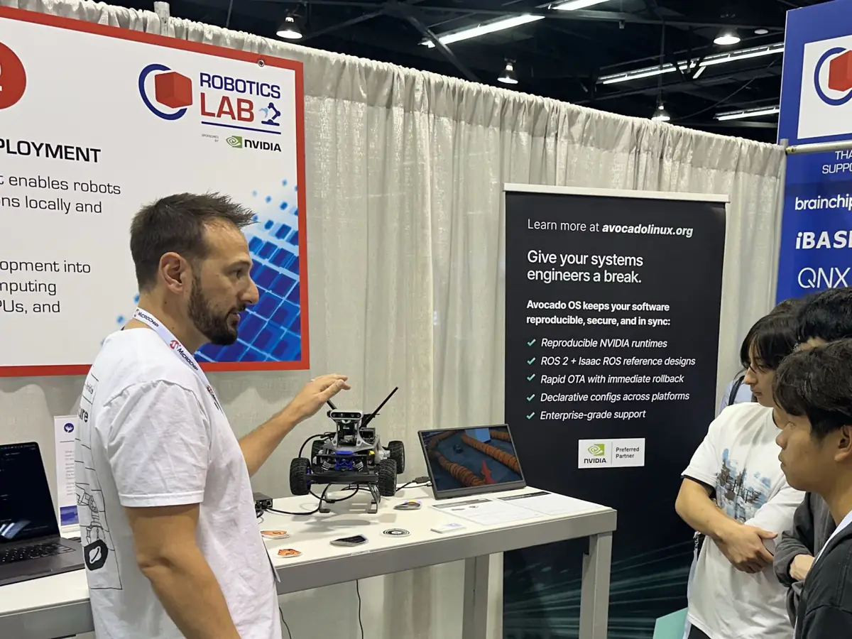 A Peridio engineer walks a group of visitors through the GOAT Racer 3 demo at the Robotics Lab booth, beside an Avocado OS banner